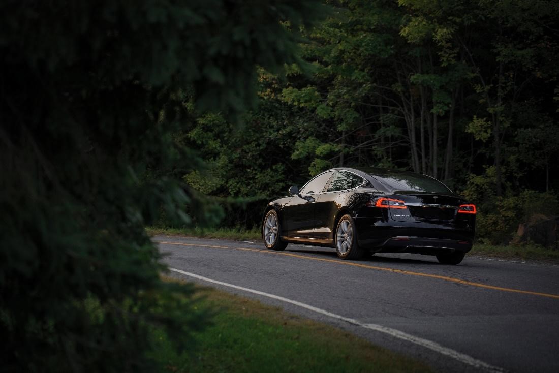 Black Tesla Model S driving on a tar road with green forest in background